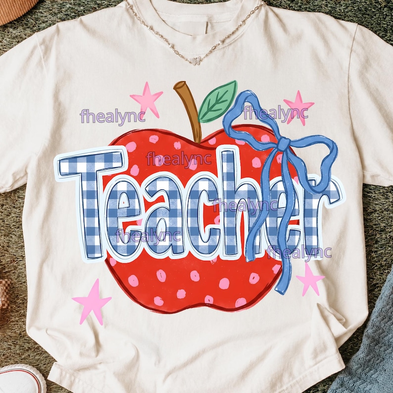 May include: A cream-colored t-shirt featuring a large red apple graphic with pink polka dots, a brown stem, and a green leaf. The word Teacher is printed in a blue and white gingham pattern across the apple, accented by a blue ribbon and pink stars.