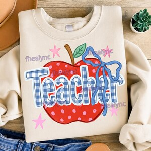 May include: A cream-colored crewneck sweatshirt featuring a large red apple graphic with a brown stem and green leaf. The word Teacher is printed in blue and white gingham letters across the apple, accented by a blue ribbon and pink stars.