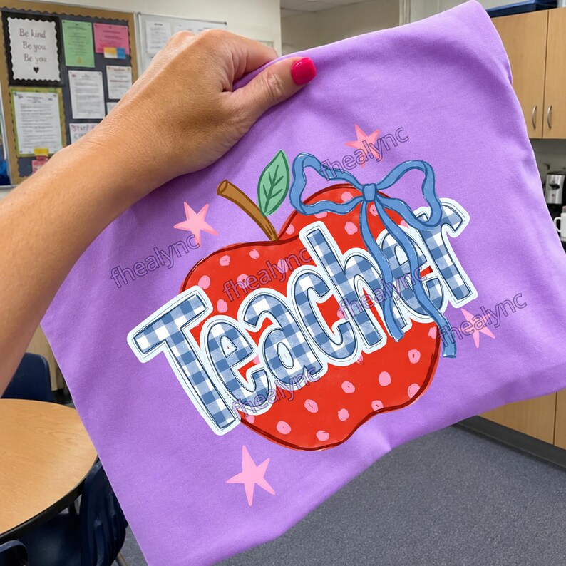 May include: A light purple t-shirt featuring a graphic of a red apple with white polka dots, a green leaf, and a blue gingham bow. The word "Teacher" is printed in large blue and white gingham letters across the apple, surrounded by pink stars.
