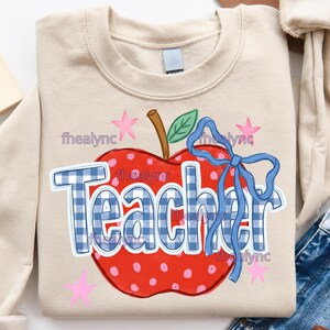 May include: A beige crewneck sweatshirt featuring a large red apple with white polka dots, a brown stem, and a green leaf. The word Teacher is printed in a blue and white gingham pattern, accented by a blue ribbon bow and small pink stars.
