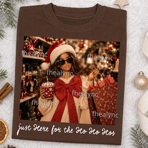 May include: A brown t-shirt featuring a graphic of a person wearing a Santa hat and sunglasses, holding a cup and a gift bag, with a large red bow on their sweater. The text "just here for the ho ho hos" is printed in white below the graphic.
