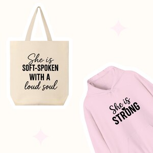 May include: A cream-colored tote bag with the text "She is soft-spoken with a loud soul" in black. Next to it is a pink hoodie with the text "She is STRONG" in black.