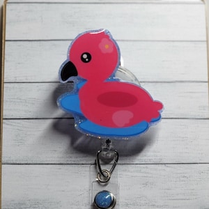Peut inclure: Un enrouleur de badge avec un flamant rose, un bec noir et un motif d'eau bleu. L'enrouleur de badge a un cordon rétractable en plastique transparent et un accent bleu et argenté. L'enrouleur de badge est fixé à un fond en bois blanc et gris.
