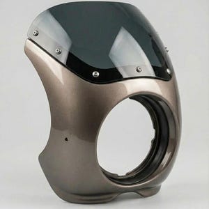 May include: A motorcycle fairing in a metallic taupe color with a dark tinted windscreen. The fairing has a circular opening and is designed to mount to the front of a motorcycle. The windscreen is secured with multiple silver rivets.