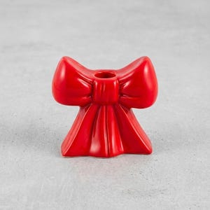 May include: A glossy red ceramic candle holder shaped like a bow. The candle holder has a circular opening at the top for a single candle. The bow design features rounded loops and a draped base, creating a decorative accent.