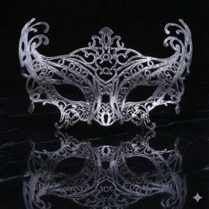 May include: A silver filigree masquerade mask with an ornate design. The mask features intricate swirls and curves, with a symmetrical pattern. The mask is displayed on a reflective surface, creating a mirrored effect. The overall aesthetic is elegant and decorative.