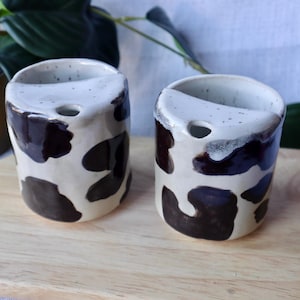 May include: Two cylindrical ceramic cups featuring a white base with irregular dark brown spots. Each cup has a distinct semi-circular rim section with a small circular opening, creating a unique silhouette. The surface has a glossy finish.