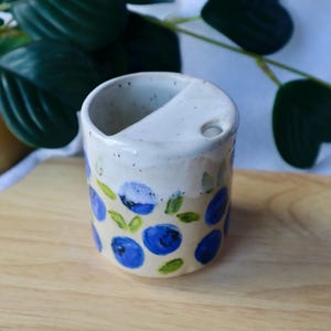 May include: A ceramic tumbler with a speckled white top section and a beige bottom section featuring a hand-painted pattern of blue blueberries and green leaves. The rim includes a built-in semi-circular ledge and a small circular opening.