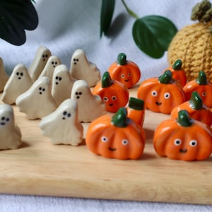 May include: A collection of small ceramic figurines featuring white ghosts with black painted eyes and orange pumpkins with green stems and smiling faces. The figures are arranged on a light wooden surface, with a crocheted pumpkin in the back.
