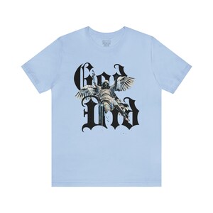 May include: Light blue t-shirt featuring a black graphic print of an angel with wings and the word "God" in a gothic font.