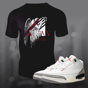 Op de afbeelding: Een zwart T-shirt met een afbeelding van een engel met vleugels en het woord "PRAY" in rode letters. De tekst "FOR MY ENEMIES" staat onder de afbeelding. Een paar witte en grijze sneakers met rode accenten.