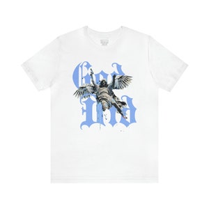 May include: White t-shirt featuring a blue graphic print of an angel with wings, flying through the air. The graphic is styled with the word "God" in large, gothic letters.