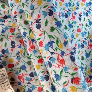 May include: Close-up of a fabric with a repeating floral pattern. The design features small flowers in red, blue, yellow, and green on a white background. The fabric is folded, showing the texture and drape of the material. A vintage tag is visible.
