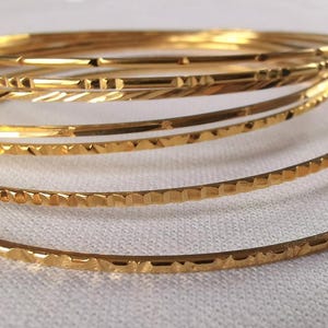 Könnte beinhalten: Eine Sammlung goldener Armreifen mit verschiedenen strukturierten Designs. Die Armreifen sind gestapelt und zeigen verschiedene Muster wie geätzte Linien und geometrische Formen. Der Schmuck wird auf einer weißen Oberfläche präsentiert.