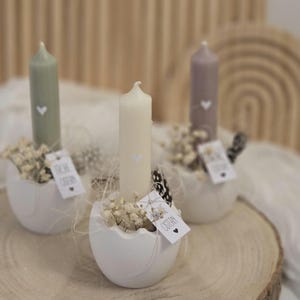 May include: Three white ceramic egg-shaped candle holders with white candles and small white flowers. Each holder has a tag that says "Frohe Ostern" in black lettering.