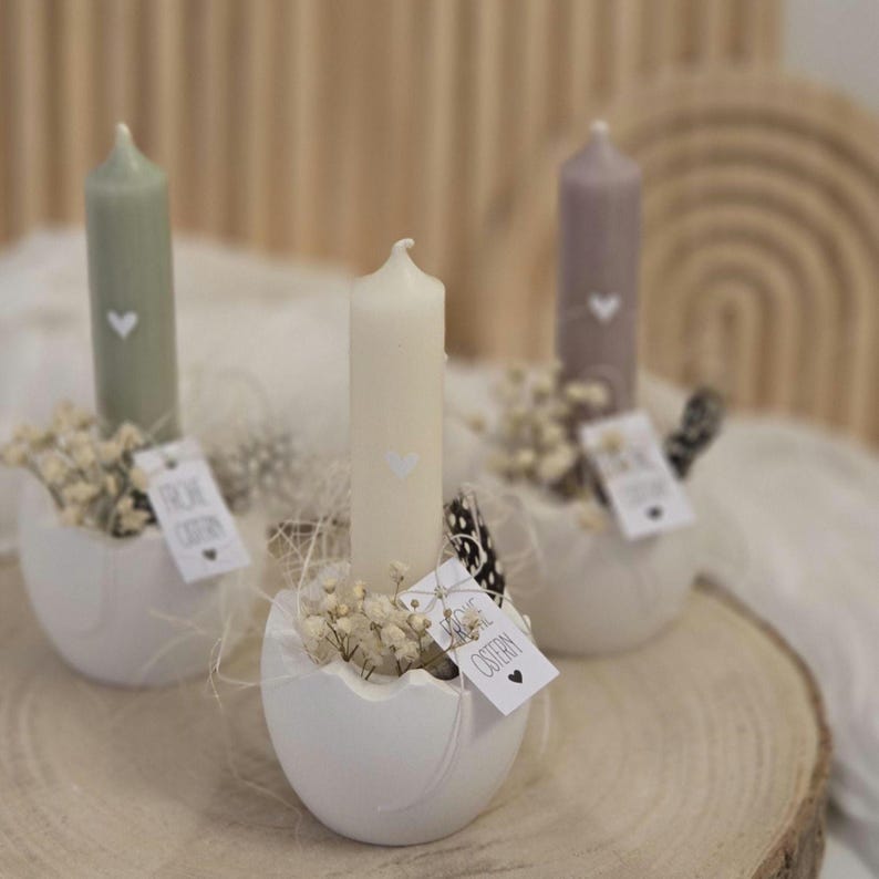 May include: Three white ceramic egg-shaped candle holders with white candles and small white flowers. Each holder has a tag that says "Frohe Ostern" in black lettering.