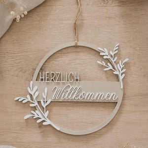Könnte beinhalten: Ein kreisförmiges Wandobjekt aus beigem Holz mit einem horizontalen Steg, der den weißen Schriftzug "HERZLICH Willkommen" trägt, sowie weißen Blattzweig-Ausschnitten an den Seiten. Das Schild ist an einer dünnen Juteschnur aufgehängt.