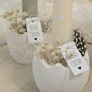 May include: Two white ceramic eggshells with a small bouquet of white baby's breath flowers in each. Each eggshell has a white tag with black text that reads "Frohe Ostern" and a small black heart.
