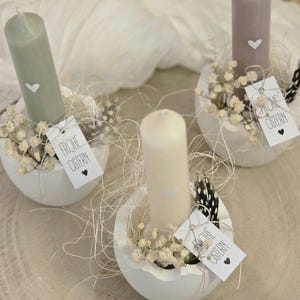 Könnte beinhalten: Drei weiße, eiförmige Kerzenhalter aus Keramik mit kleinen weißen Kerzen. Jeder Halter ist mit weißen Babyschnabelblumen, einem kleinen weißen Herzen und einer schwarz-weißen Feder verziert. Jeder Halter hat ein kleines Schild mit der Aufschrift "Frohe Ostern" in schwarzer Schrift.