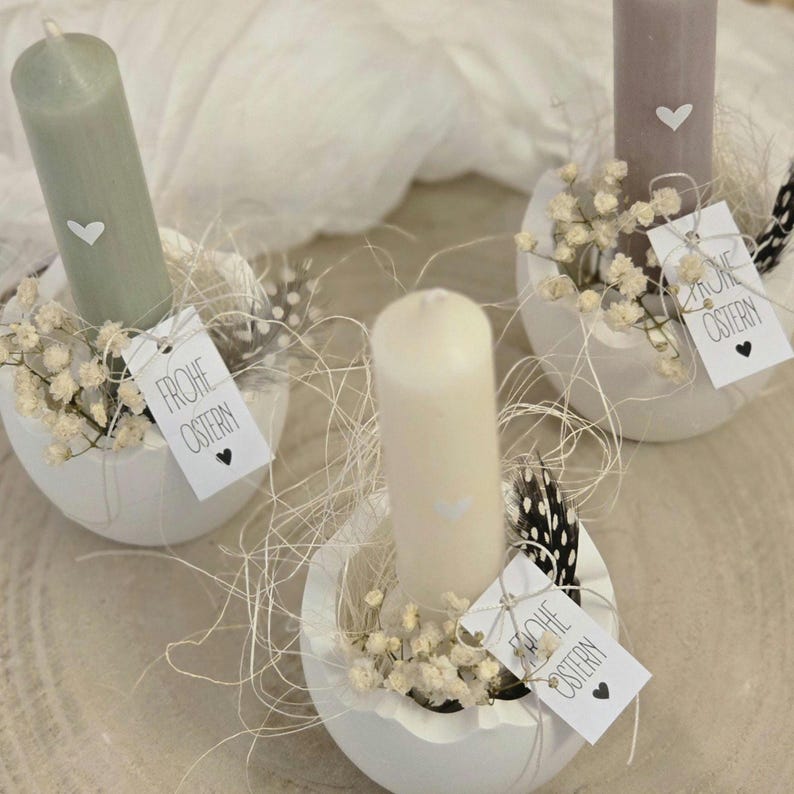 May include: Three white ceramic egg-shaped candle holders with small white candles. Each holder is decorated with white baby's breath flowers, a small white heart, and a black and white feather. Each holder has a small tag that says "Frohe Ostern" in black lettering.