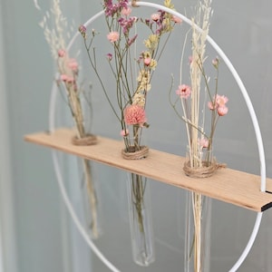 以下が含まれることがあります： 白い金属製の輪に木製の棚と、乾燥した花で満たされたガラス製の試験管が3本あります。花はピンク、白、ベージュです。