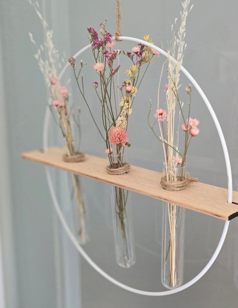 以下が含まれることがあります： 白い金属製の輪に木製の棚と、乾燥した花で満たされたガラス製の試験管が3本あります。花はピンク、白、ベージュです。