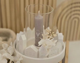May include: White ceramic candle holder with a glass cylinder and a grey candle. The candle holder is decorated with small white houses and a white flower. The candle holder is on a wooden surface.