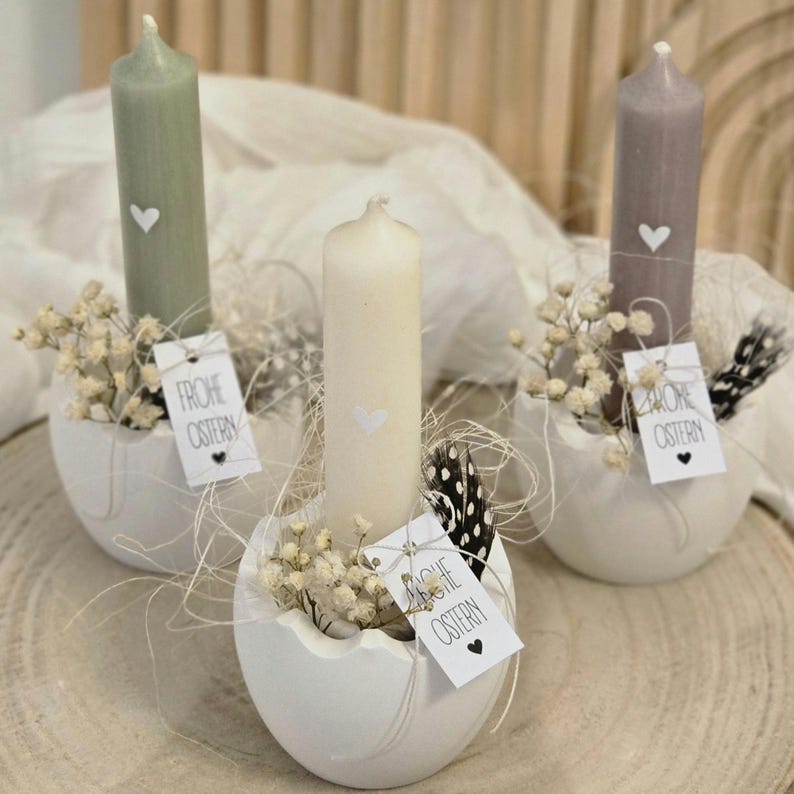 May include: Three white ceramic egg-shaped candle holders with small white candles and dried white flowers. Each holder has a small tag with the text "Frohe Ostern" in black.