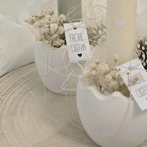 May include: Two white ceramic eggshells with a small bouquet of white baby's breath flowers in each. The eggshells are sitting on a wooden surface. Each eggshell has a small white tag with black text that says "Frohe Ostern" which means "Happy Easter" in German.