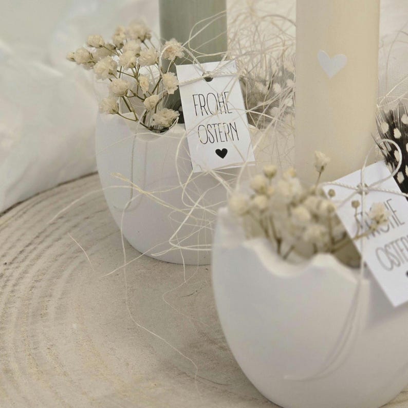 May include: Two white ceramic eggshells with a small bouquet of white baby's breath flowers in each. The eggshells are sitting on a wooden surface. Each eggshell has a small white tag with black text that says "Frohe Ostern" which means "Happy Easter" in German.