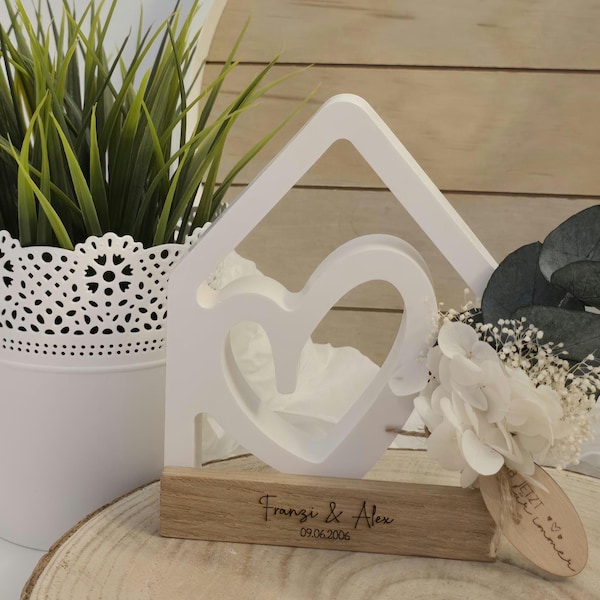 May include: A white wooden house-shaped decoration with a heart cutout, resting on a wooden base. The base is engraved with a name and date. A small bouquet of white flowers and greenery is attached.