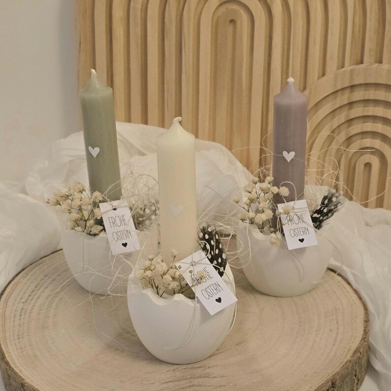 May include: Three white ceramic egg-shaped candle holders with white candles and small white flowers. Each holder has a tag with the text "Frohe Ostern" in black.