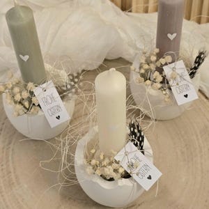 May include: Three white egg-shaped candle holders with white candles and small white flowers. Each holder has a tag that says "Frohe Ostern" which means "Happy Easter" in German.