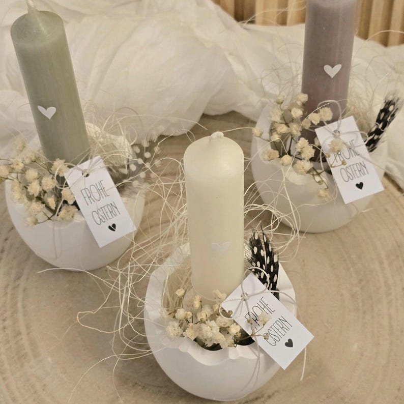 May include: Three white egg-shaped candle holders with white candles and small white flowers. Each holder has a tag that says "Frohe Ostern" which means "Happy Easter" in German.