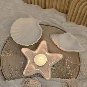 Könnte beinhalten: Dekorative Elemente im Küstenstil. Ein sternförmiger Kerzenhalter mit brennender Kerze, zwei Muschelschalen und eine Muschel. Die Artikel sind weiß und beige, angeordnet auf einer Holzscheibe und weißem Stoff. Der Kerzenhalter hat einen Durchmesser von ca. 10 cm.