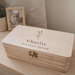 May include: A light-coloured wooden box with a hinged lid and a metal clasp. The box is engraved with the name "Charlie" and the phrase "forever loved." A framed photo and a potted plant are in the background.
