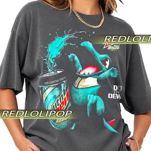 Pode incluir: Uma t-shirt cinzenta escura de manga curta com um gráfico de uma criatura azul-esverdeada ao lado de uma lata de Mountain Dew Baja Blast com um salpico de líquido. O texto diz Baja Blast, For A Limited Time e Do The Dew.