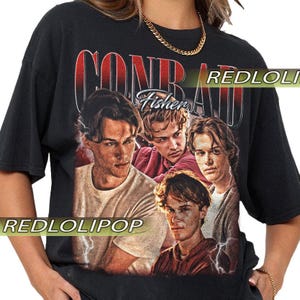 Pode incluir: Camiseta preta com um gráfico de quatro jovens e o texto "CONRAD FISHER" em vermelho e branco. A camiseta tem um visual vintage e desgastado, com o nome da marca "REDLOLIPOP" impresso na frente.