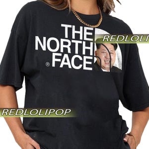 May include: Black short-sleeved t-shirt with the text "THE NORTH FACE" in white. The image of a person is incorporated into the design. The shirt has a gold chain necklace.