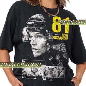 Könnte beinhalten: Schwarzes T-Shirt mit einem verblassten Grafikdruck eines Eishockeyspielergesichts, Helm und der gelben Nummer 81. Der Name "ILYA ROZANOV" ist darunter gedruckt. Das Shirt hat einen Vintage-Look.