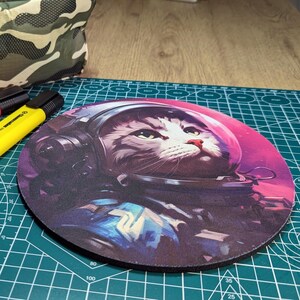 Puede incluir: Un ratón de ordenador redondo con una ilustración colorida de un gato con un casco y un traje de astronauta. El gato mira hacia la derecha y el fondo es una nebulosa rosa y morada.