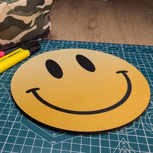 May include: Yellow smiley face mouse mat with a black outline. The mouse mat is on a green cutting mat.