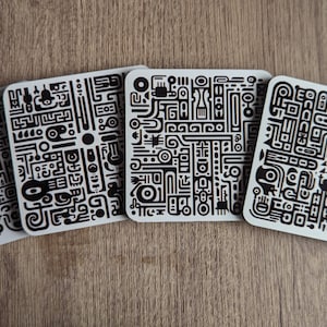 Puede incluir: Cuatro posavasos cuadrados con un patrón geométrico en blanco y negro. Los posavasos están hechos de un material claro y tienen una superficie lisa.