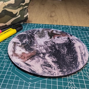 May include: A round black mouse mat with a detailed image of Earth, showing clouds and landmasses. The mouse mat is on a green cutting mat.