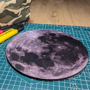 以下が含まれることがあります： 月の表面の詳細な画像が描かれた、丸い黒いマウスパッド。マウスパッドは緑色のカッティングマットの上に置かれています。