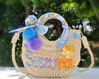 May include: A straw tote bag with a blue and white floral print fabric handle. The bag has a half-circle shape and is decorated with pom-poms in blue and purple, a yellow flower, and the name "Suzy Rae" in colorful embroidery.