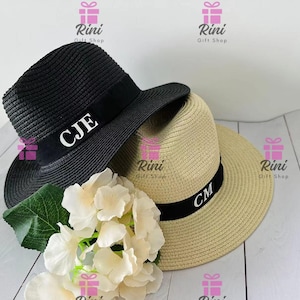 Könnte beinhalten: Zwei Strohhüte mit schwarzen Bändern und weißen Initialen. Einer ist schwarz mit "CJE" auf dem Band, der andere ist beige mit "CM". Die Hüte liegen neben weißen Blumen und grünen Blättern.