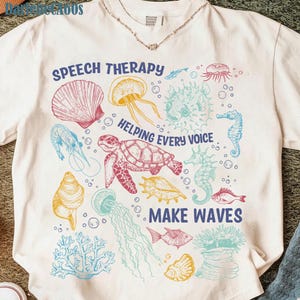 May include: Cream-colored t-shirt with a colorful sea life design. The shirt features illustrations of seashells, jellyfish, seahorses, and a turtle. Text on the shirt reads "SPEECH THERAPY HELPING EVERY VOICE MAKE WAVES."