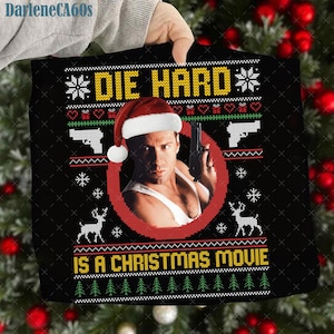 May include: Black Christmas-themed graphic tee with the text "DIE HARD IS A CHRISTMAS MOVIE" in yellow and green. The design includes a portrait of a man wearing a Santa hat, holding a gun, and festive elements.