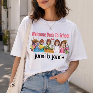 May include: A white short-sleeve t-shirt featuring a graphic print with the text "Welcome Back To School" and "junie b. jones" in colorful lettering. The illustration depicts five cartoon characters with glasses and various school supplies.